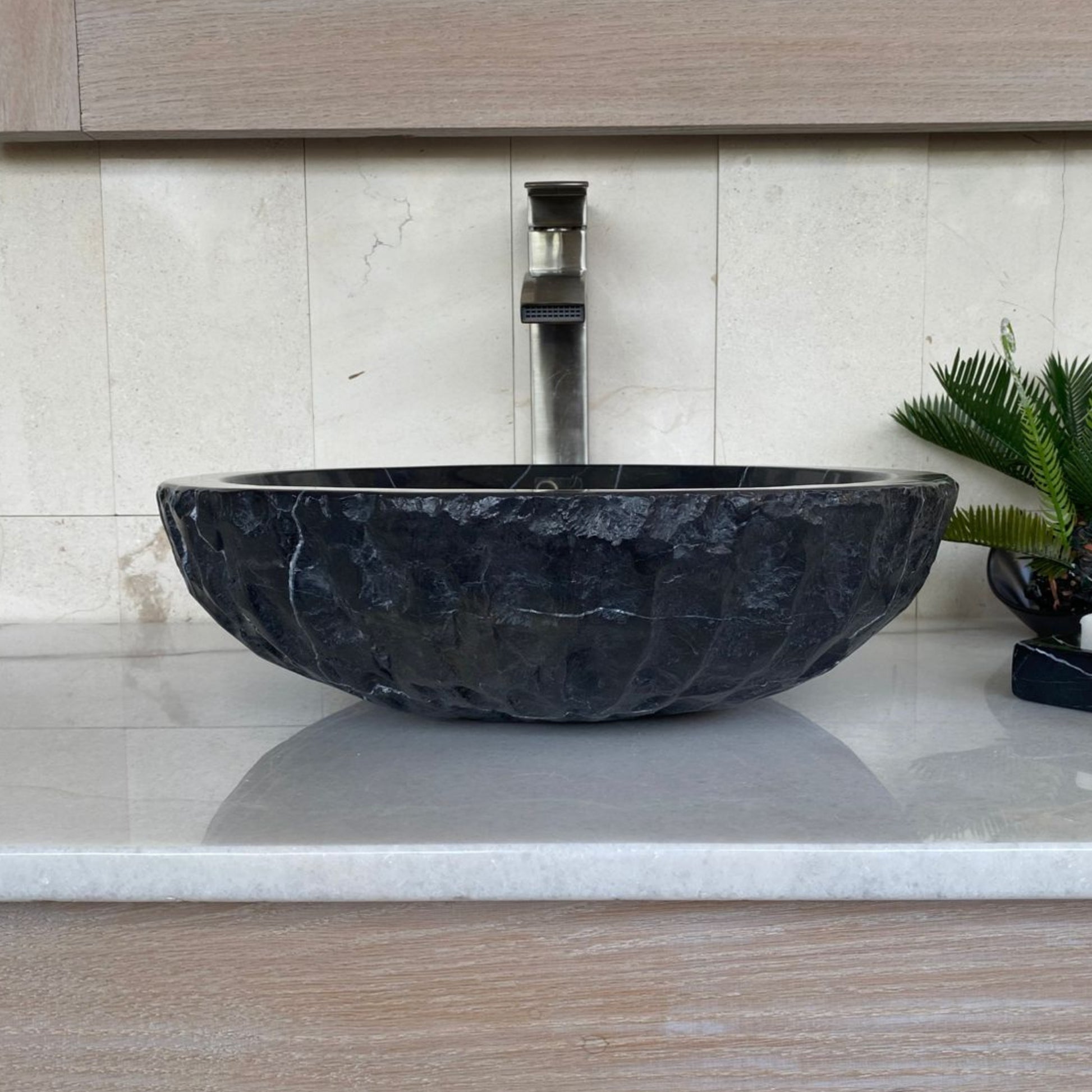 Handcrafted black marble round bowl vessel sink with chiseled exterior, polished interior, and natural white veining displayed on a modern bathroom vanity countertop.