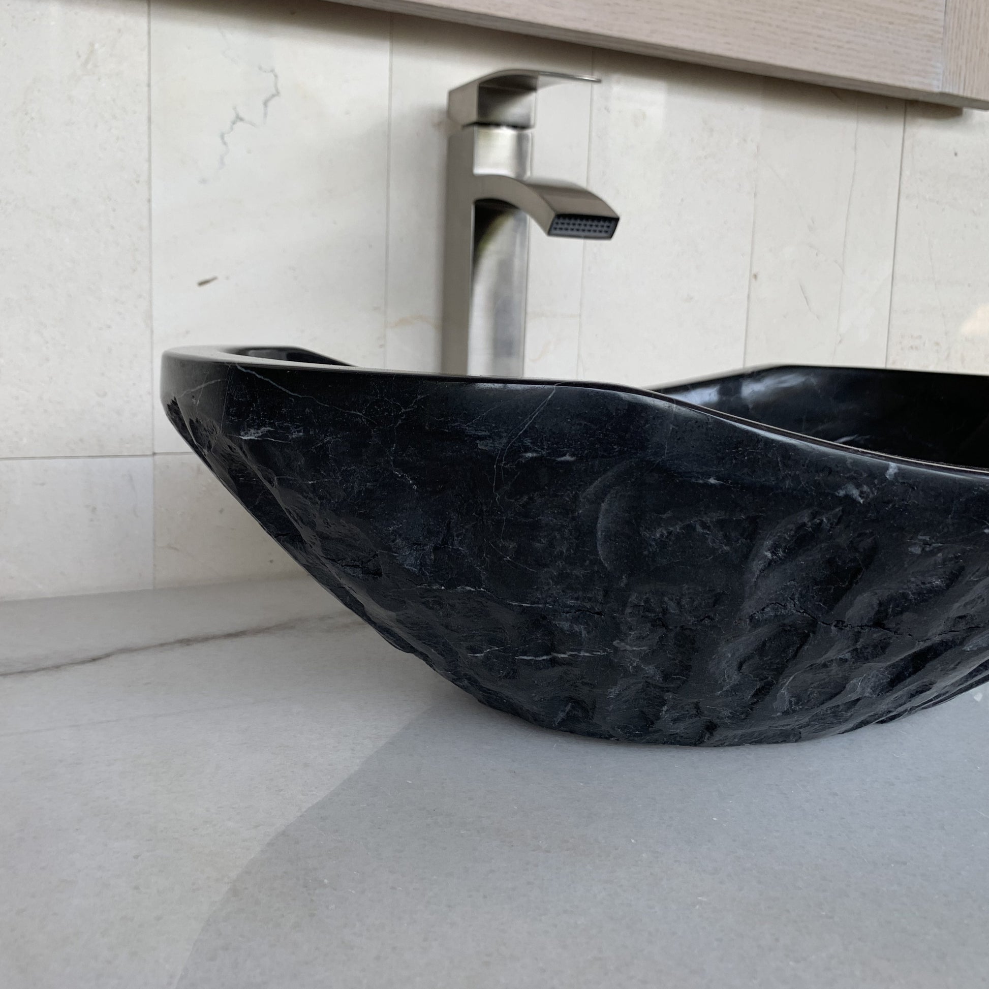 Side angle view of handcrafted black marble oval vessel sink featuring sculpted chiseled natural stone exterior, elegant curved silhouette, polished interior basin, and luxury modern bathroom vanity styling.