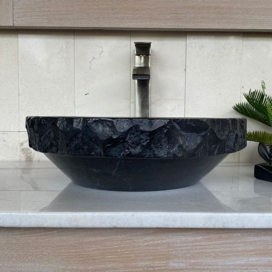 Handcrafted black marble round vessel sink with polished interior, sculpted chiseled exterior, and natural white veining displayed on a modern bathroom vanity countertop.