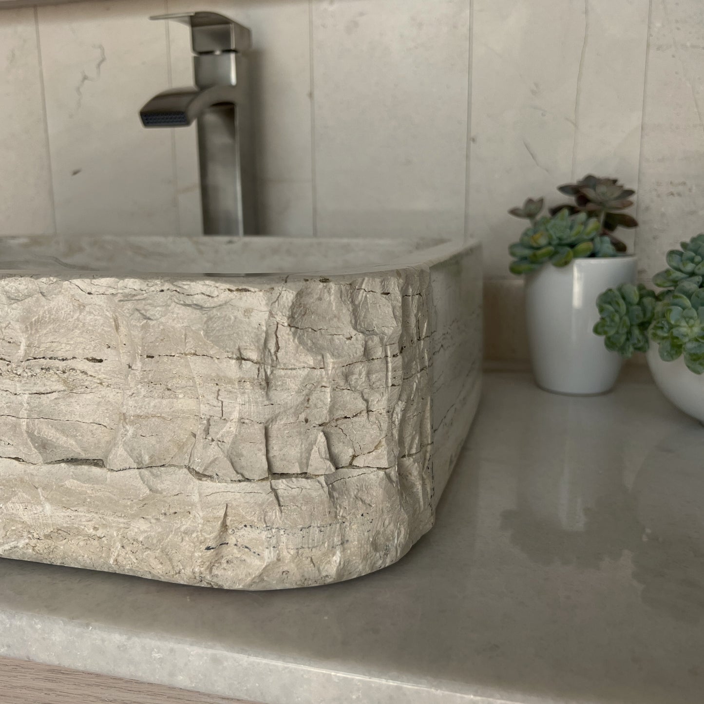 Side-angle view of a rectangular chiseled tan travertine vessel sink, showcasing the split texturing where the rugged hand-carved stone corner transitions into a smooth polished side wall.