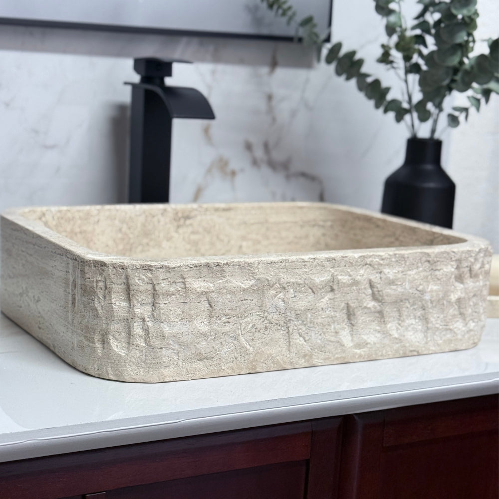 Side-angle view of a rectangular chiseled tan travertine vessel sink, showcasing the rugged hand-carved stone exterior, paired with a polished interior bowl.
