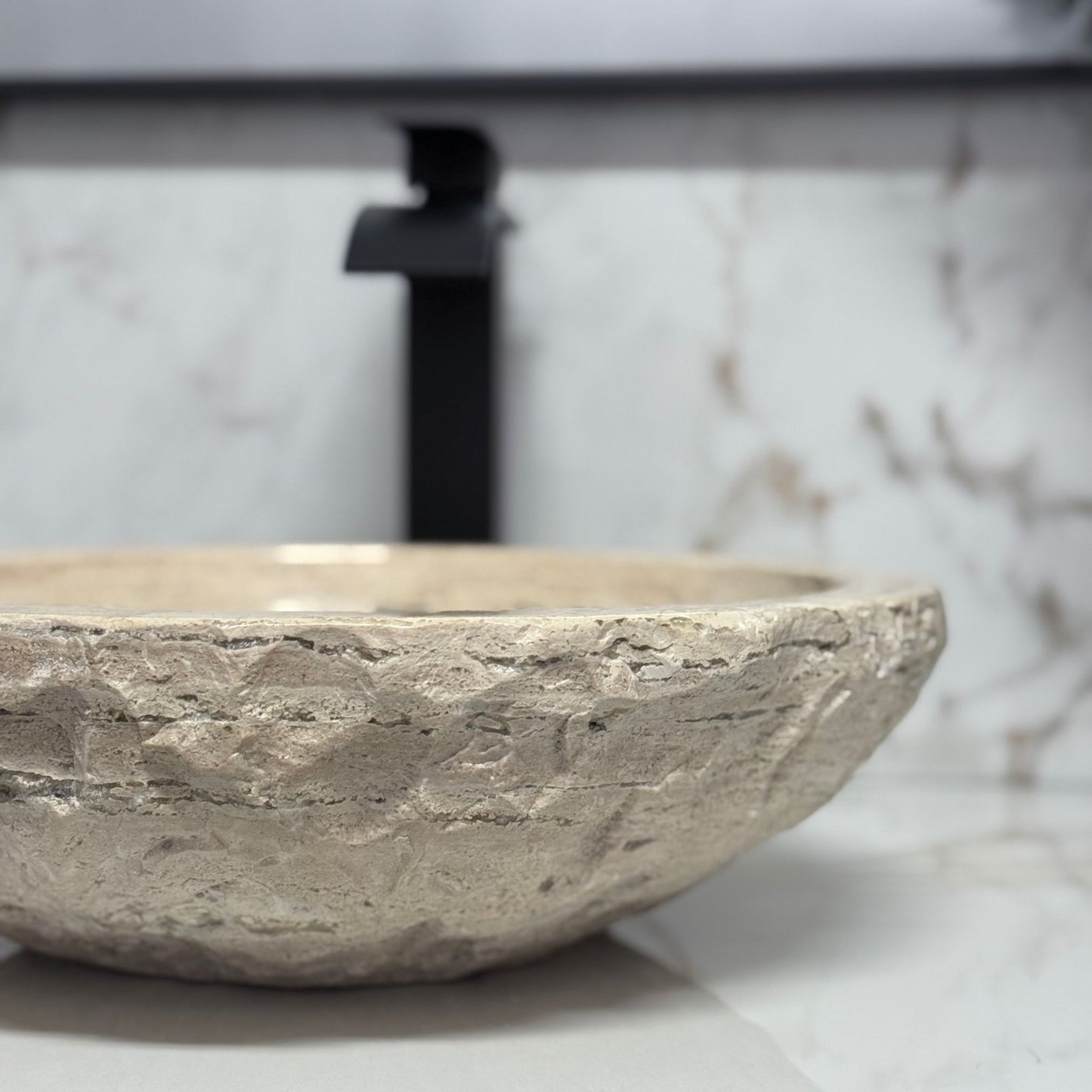 Close-up of a round beige travertine vessel sink highlighting the detailed hand-carved chiseled exterior and smooth polished interior, styled with a blurred matte black faucet background.