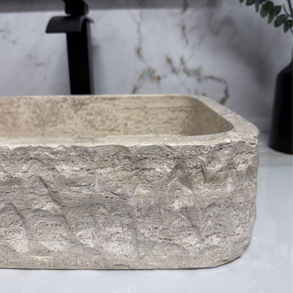 Detailed profile view of a rectangular beige travertine bathroom basin, showcasing the heavy chiseled rustic finish on its outer walls and the rounded corner geometry.
