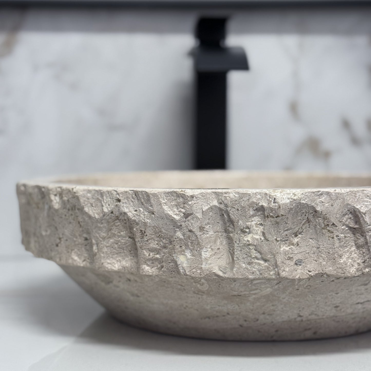 Close-up side view of a round beige travertine vessel sink, showcasing the sharp contrast between the hand-carved chiseled upper rim band and the smooth tapered lower exterior.