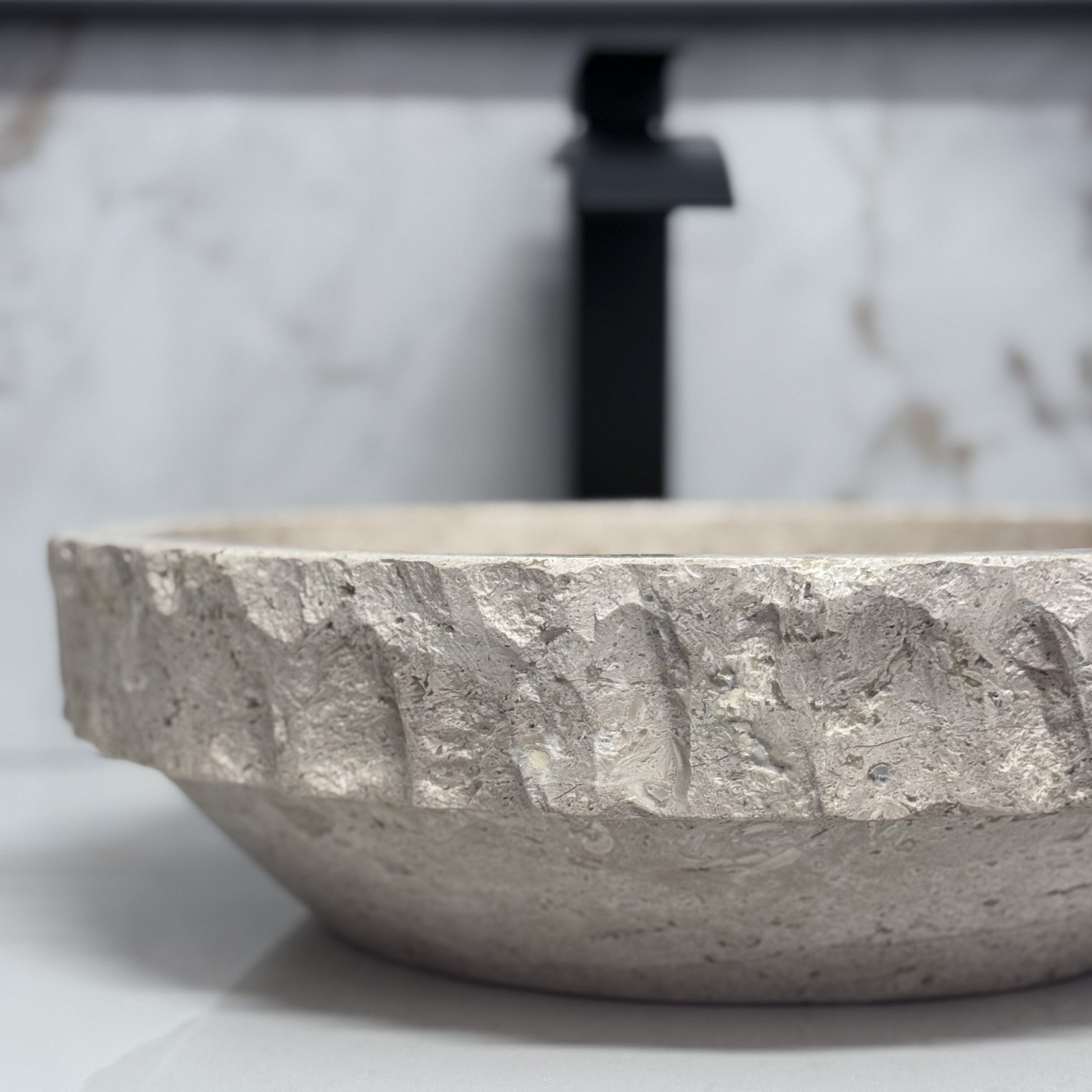 Close-up side view of a round beige travertine vessel sink, showcasing the sharp contrast between the hand-carved chiseled upper rim band and the smooth tapered lower exterior.