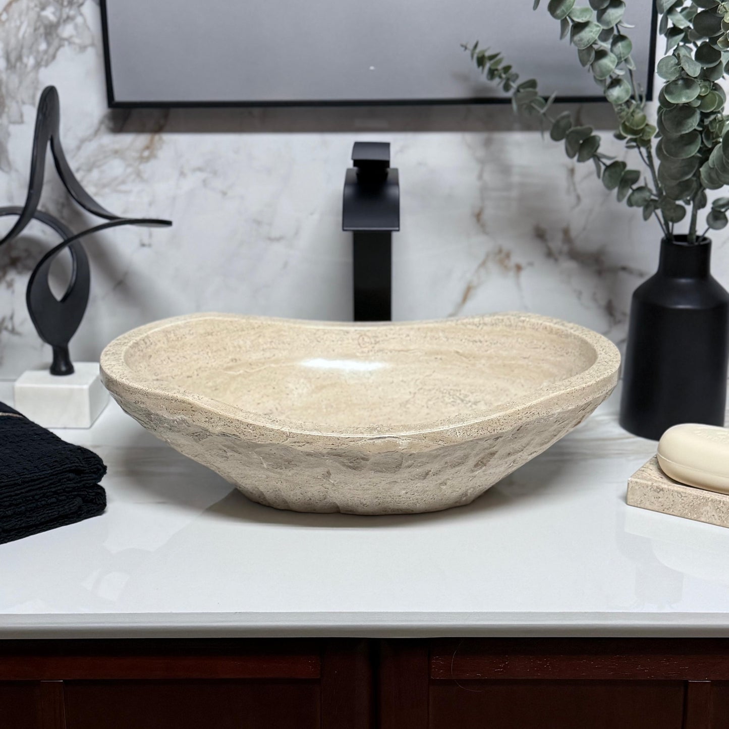 Symmetrical oval bathroom wash basin made of beige travertine stone, featuring a heavily chiseled rustic exterior surface and a curved rim profile for above-counter placement.