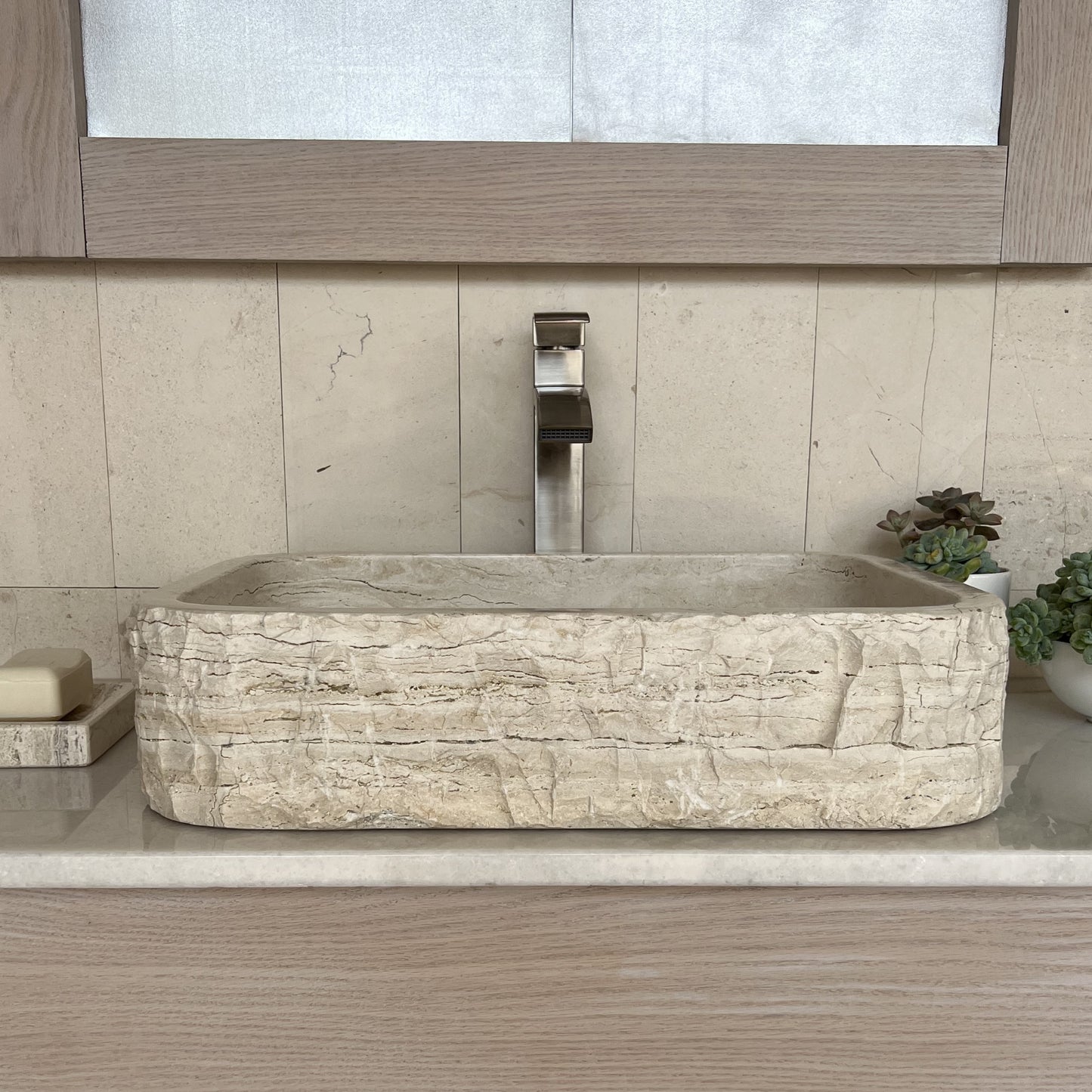 Straight-on view of a rectangular natural stone bathroom basin made of beige travertine, highlighting the heavy chiseled rustic finish on its front wall, styled with a modern silver faucet and matching stone tile.