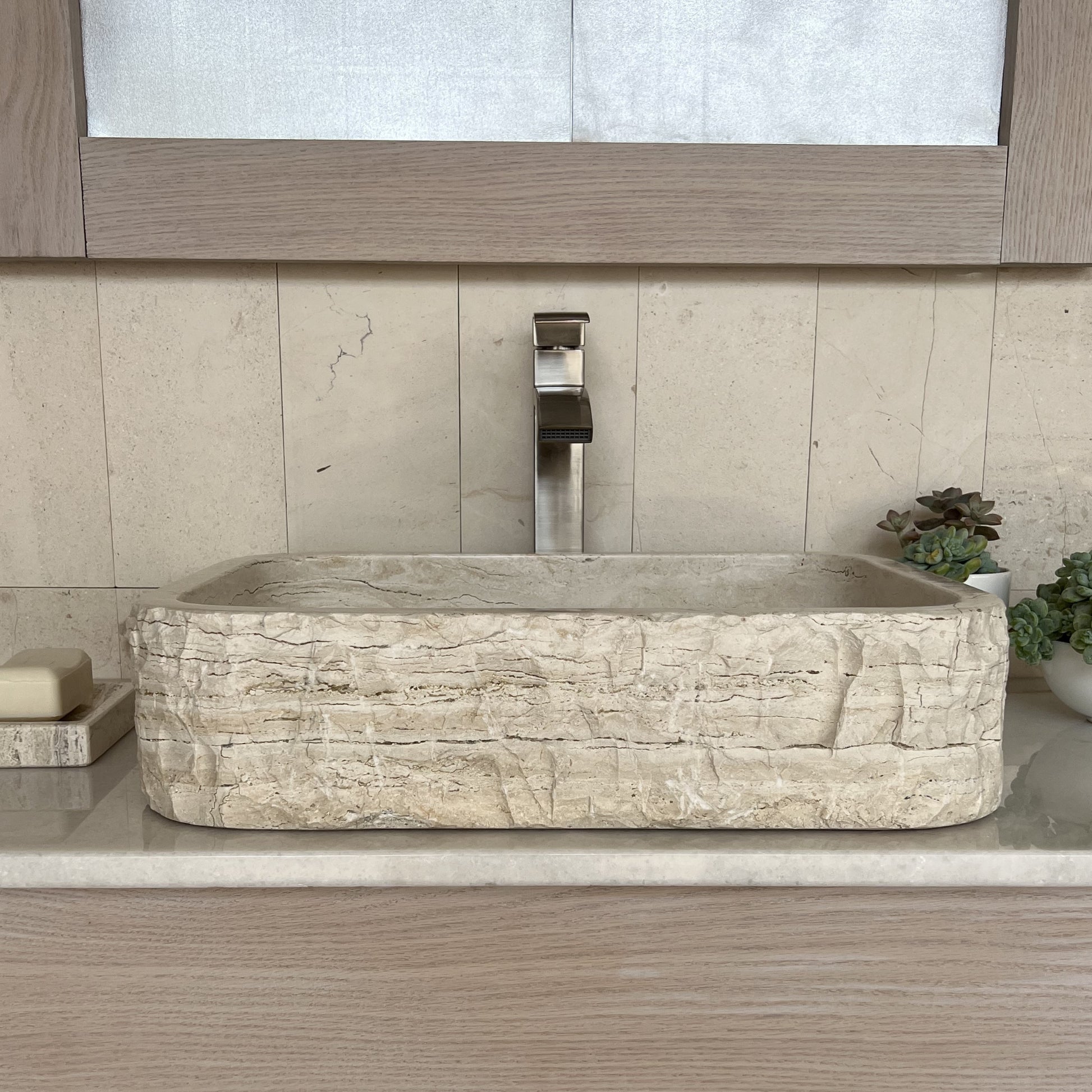 Straight-on view of a rectangular natural stone bathroom basin made of beige travertine, highlighting the heavy chiseled rustic finish on its front wall, styled with a modern silver faucet and matching stone tile.