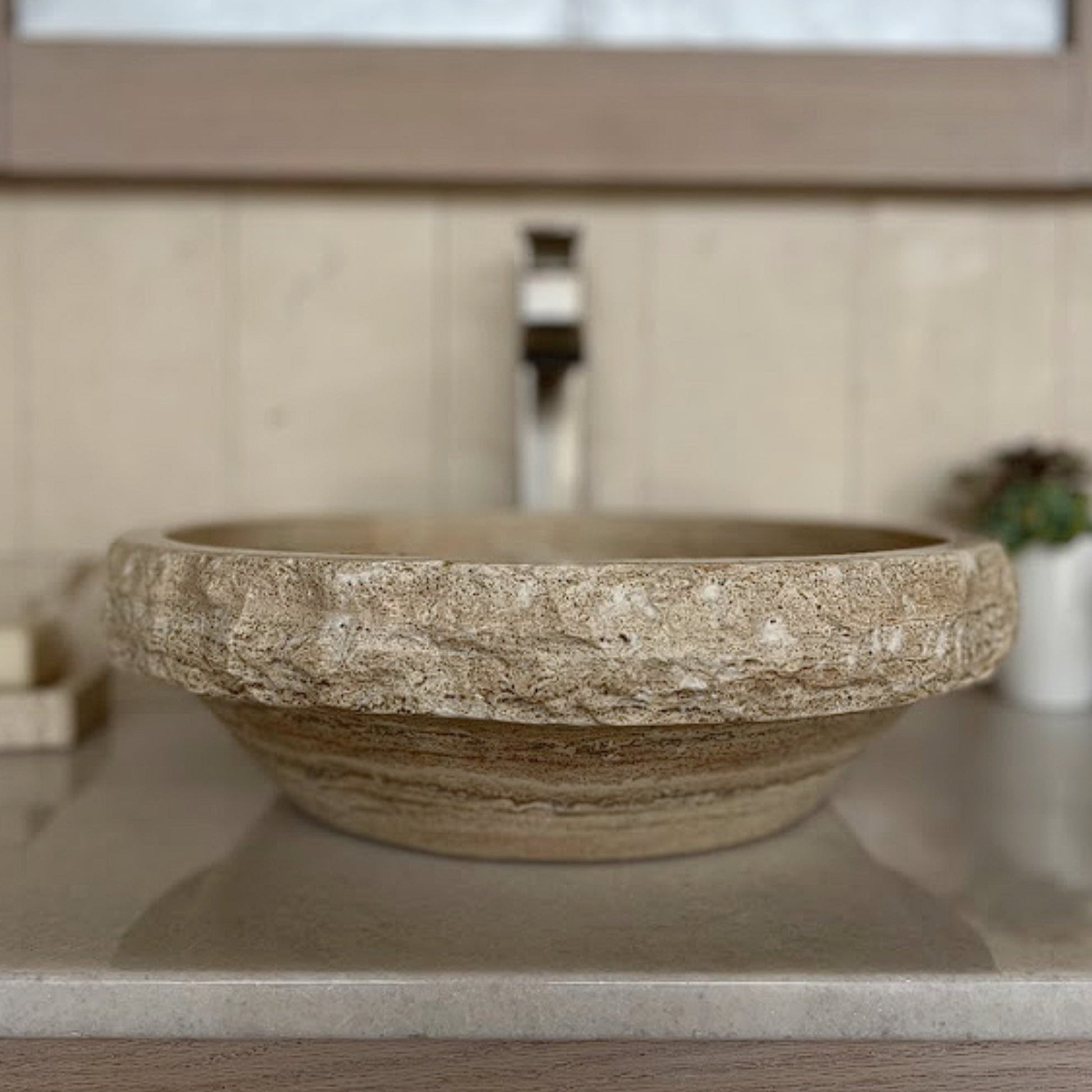 Front eye-level view of a round beige travertine vessel sink showcasing the natural stone texture contrast between the rustic chiseled upper ring and the smooth polished lower exterior, styled on a polished vanity countertop with a brushed nickel faucet backdrop.
