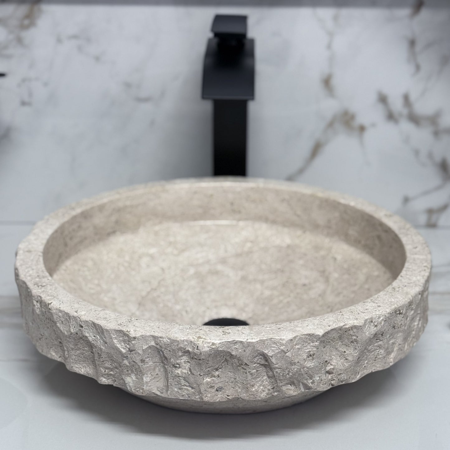 High-angle view looking into a round beige travertine vessel sink, showcasing the smooth polished interior basin contrasted by a thick, hand-carved chiseled upper rim band, styled with a modern black faucet background.