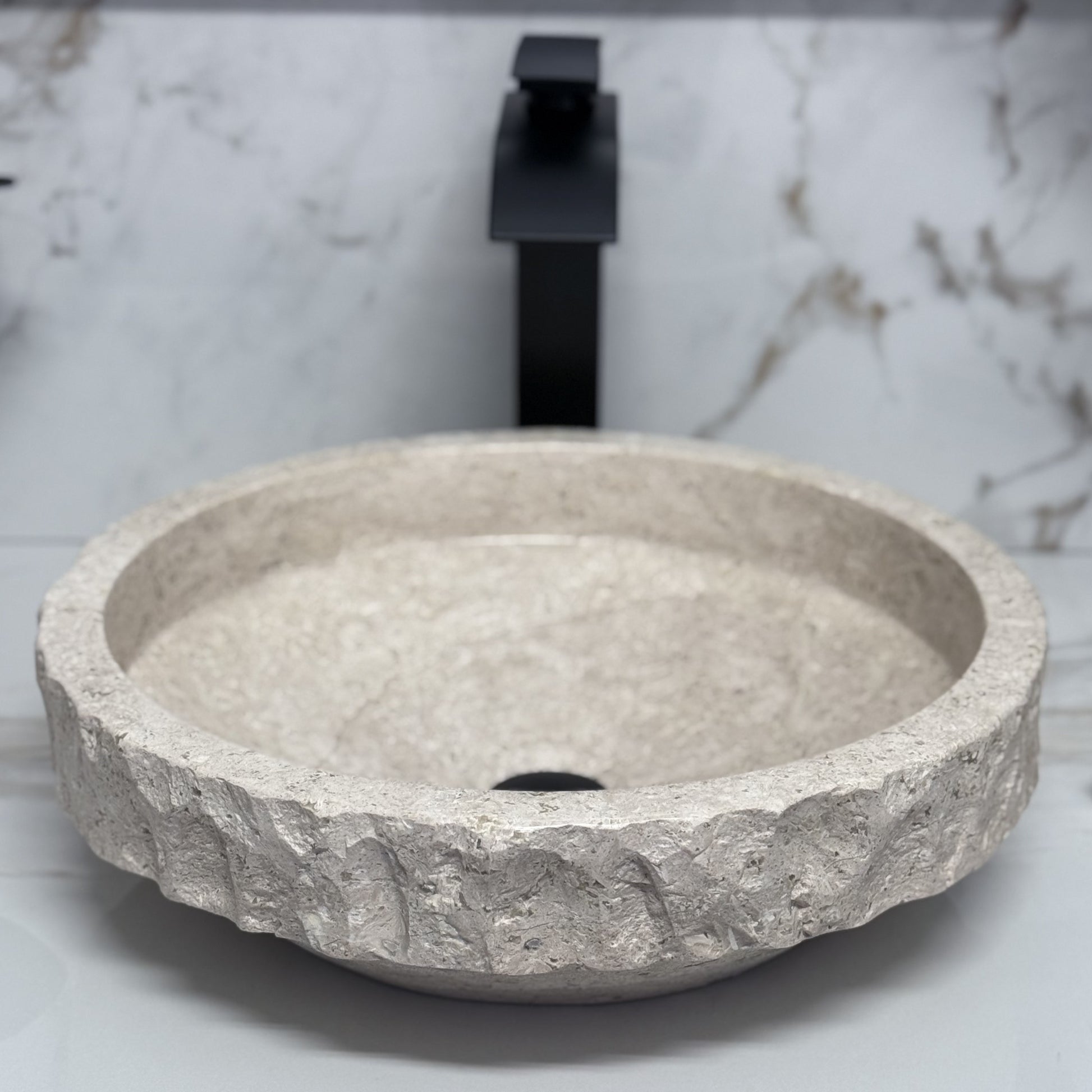 High-angle view looking into a round beige travertine vessel sink, showcasing the smooth polished interior basin contrasted by a thick, hand-carved chiseled upper rim band, styled with a modern black faucet background.