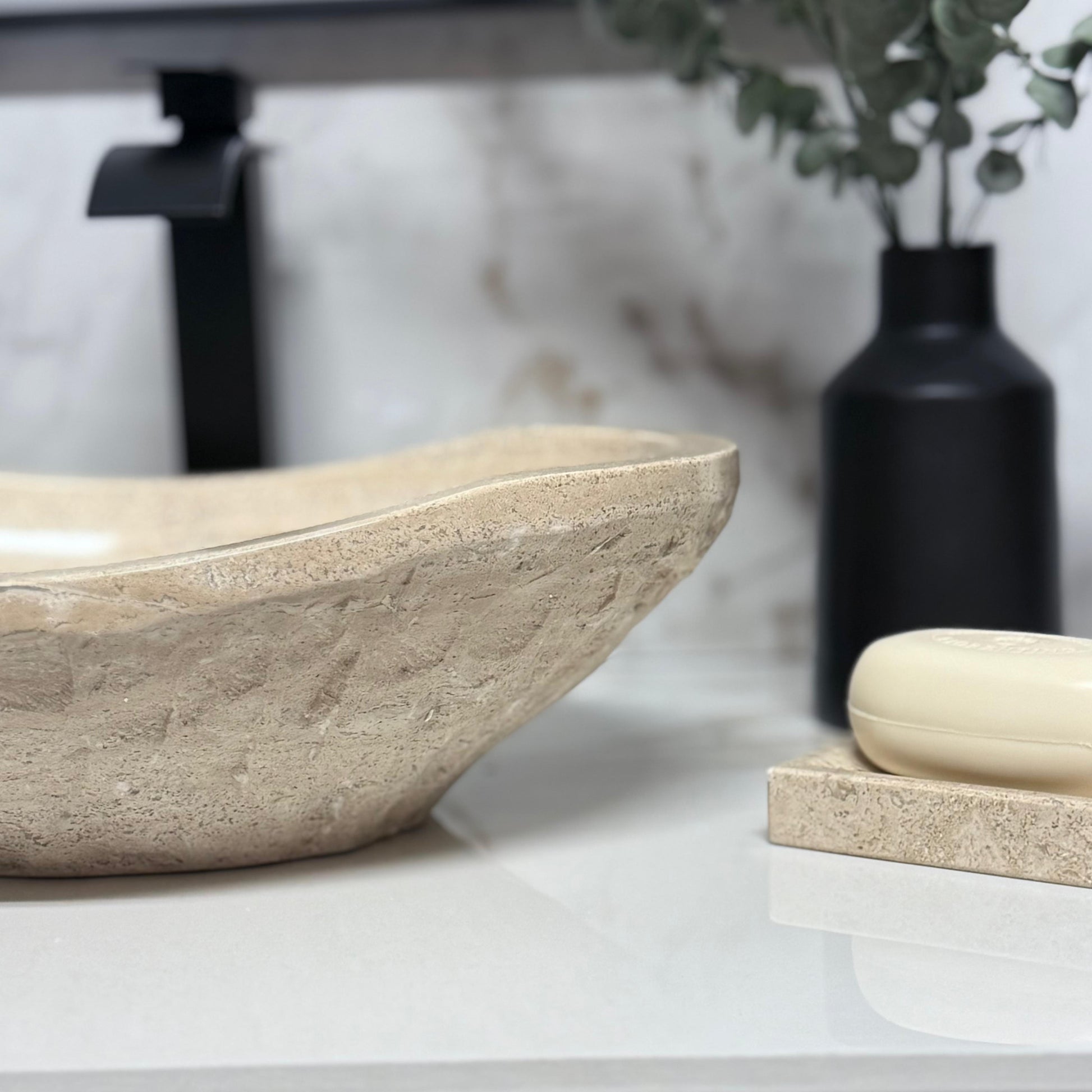 Close-up side view of an oval chiseled tan travertine vessel sink showcasing the rugged, hand-carved stone exterior texture and smooth rim, paired with a matching natural stone travertine soap dish.