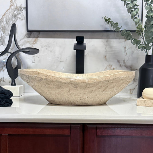 Front eye-level view of an oval chiseled tan travertine vessel sink, showcasing its rugged hand-carved stone exterior texture and distinctive curved rim profile for above-counter installation.