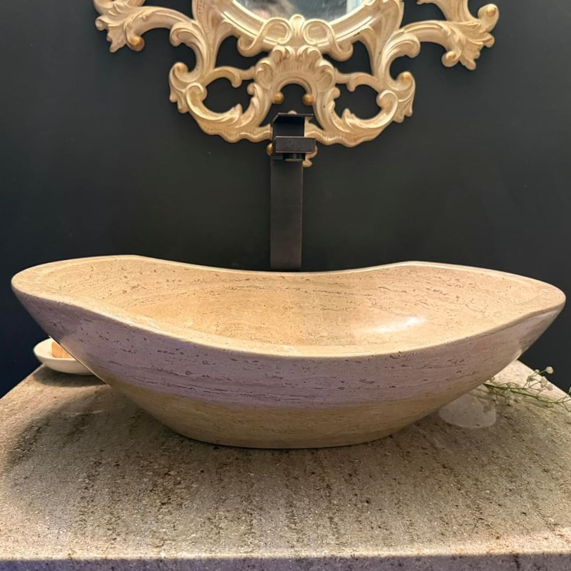Modern oval vessel sink made of fully polished travertine stone, shown in a luxury bathroom vanity setting with an elegant gold wall mirror backdrop.