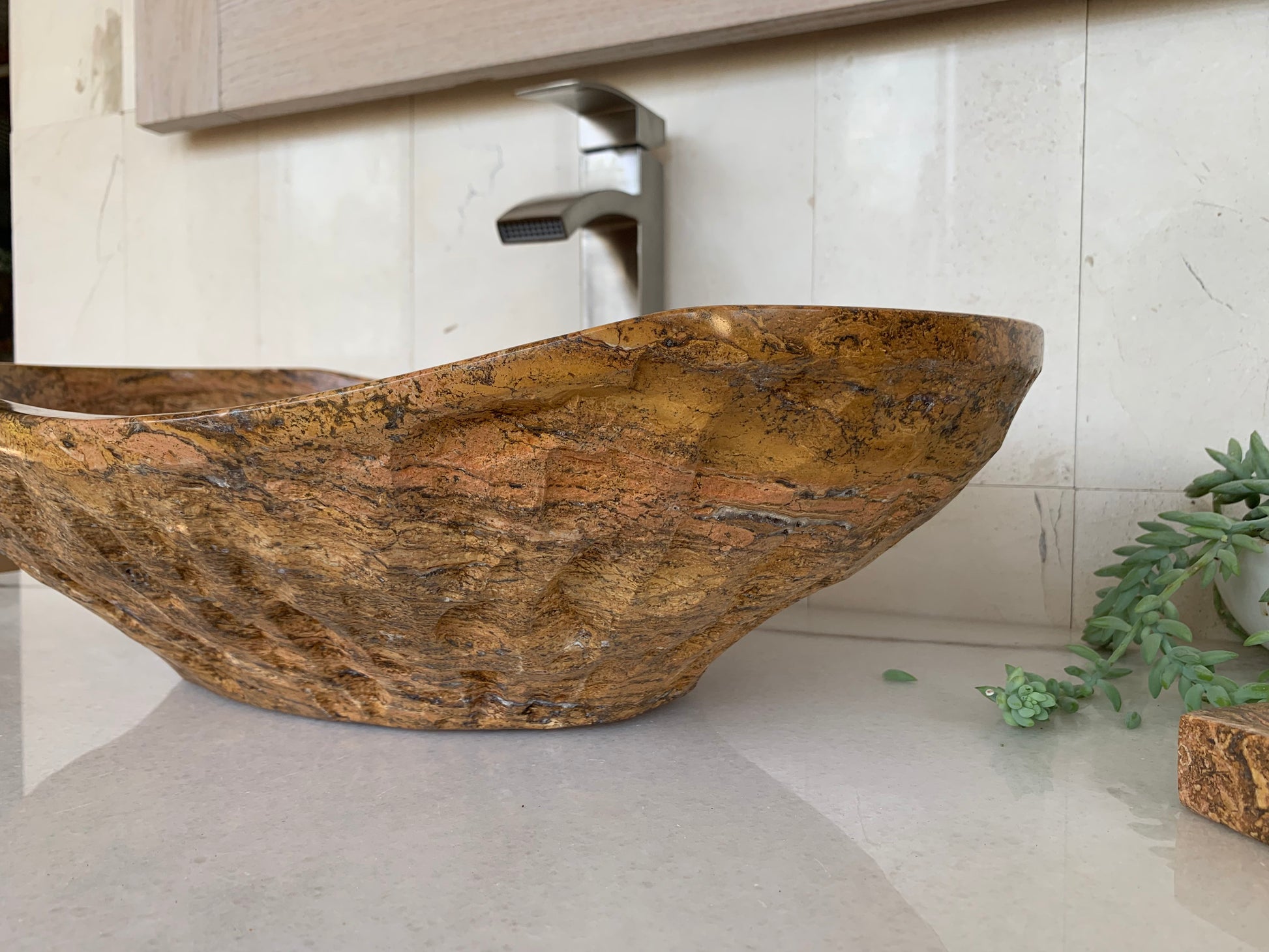 Side view of a handcrafted oval red travertine vessel sink with natural textured stone exterior on a modern bathroom countertop.