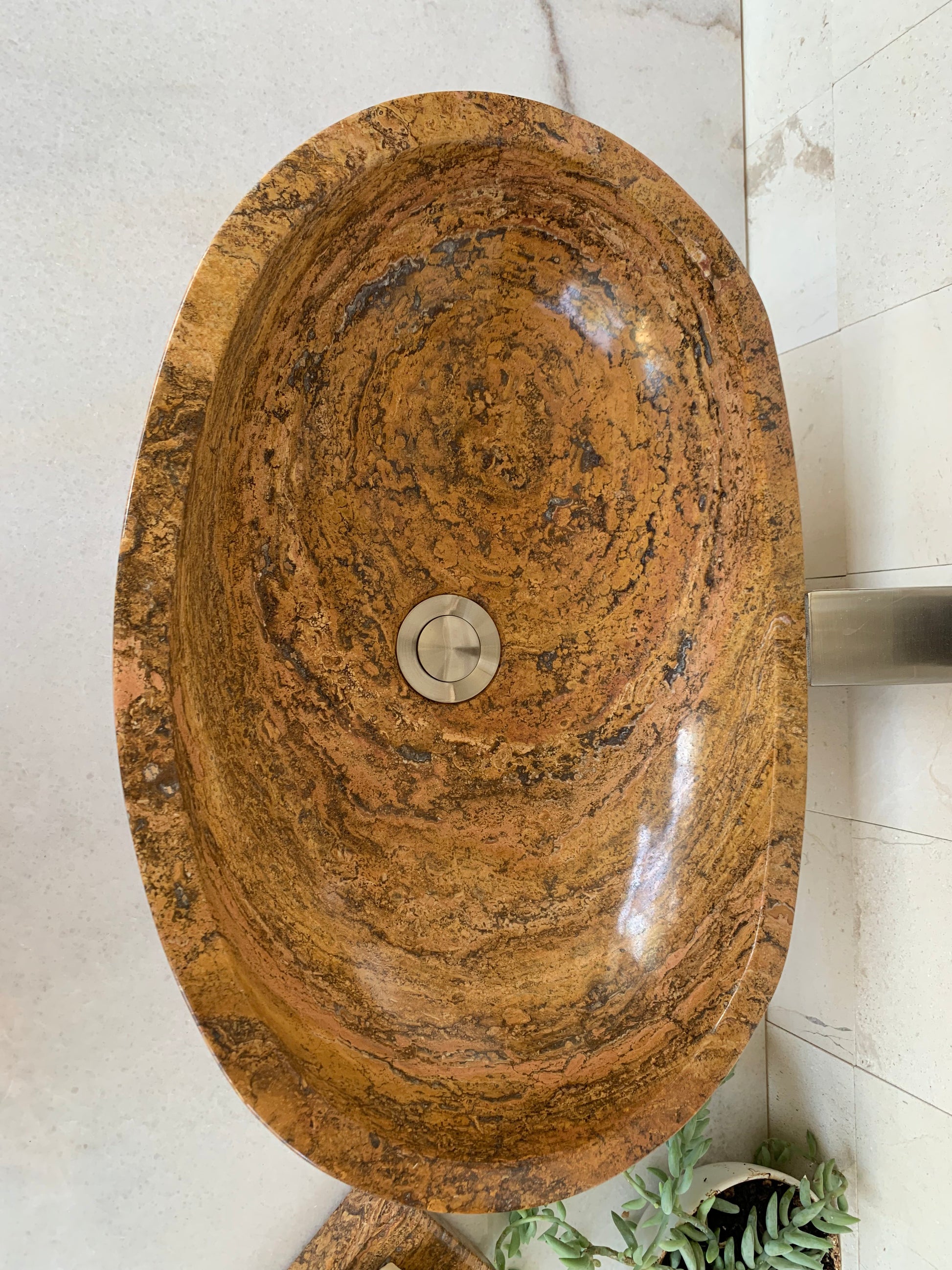 Top view of a handcrafted oval red travertine vessel sink with polished natural stone interior and drain on a modern bathroom countertop.