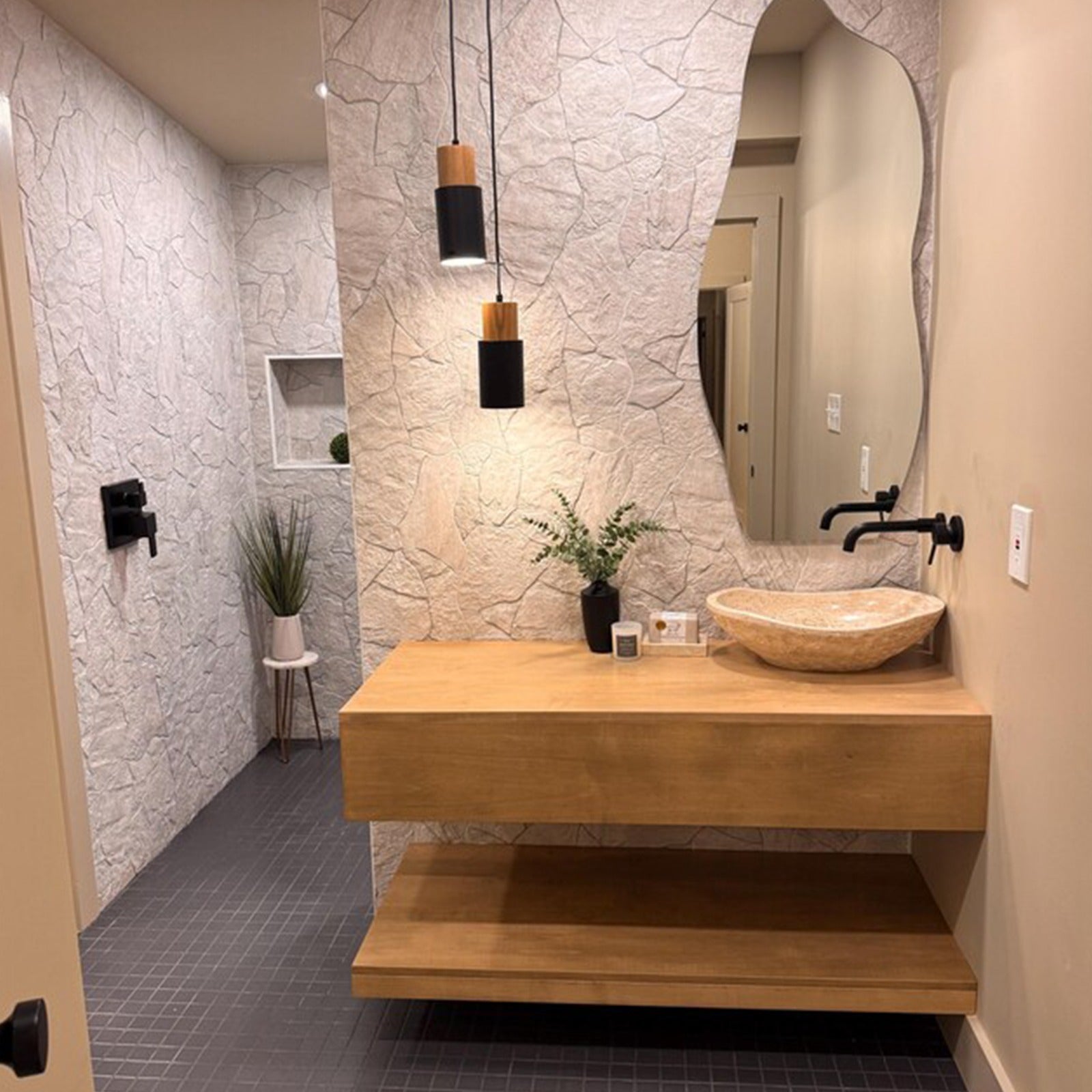 Hand-carved oval travertine bathroom wash basin with a curved silhouette, displayed in a designer bathroom setting featuring a wooden counter, black wall-mounted faucet, and an organic shaped mirror.