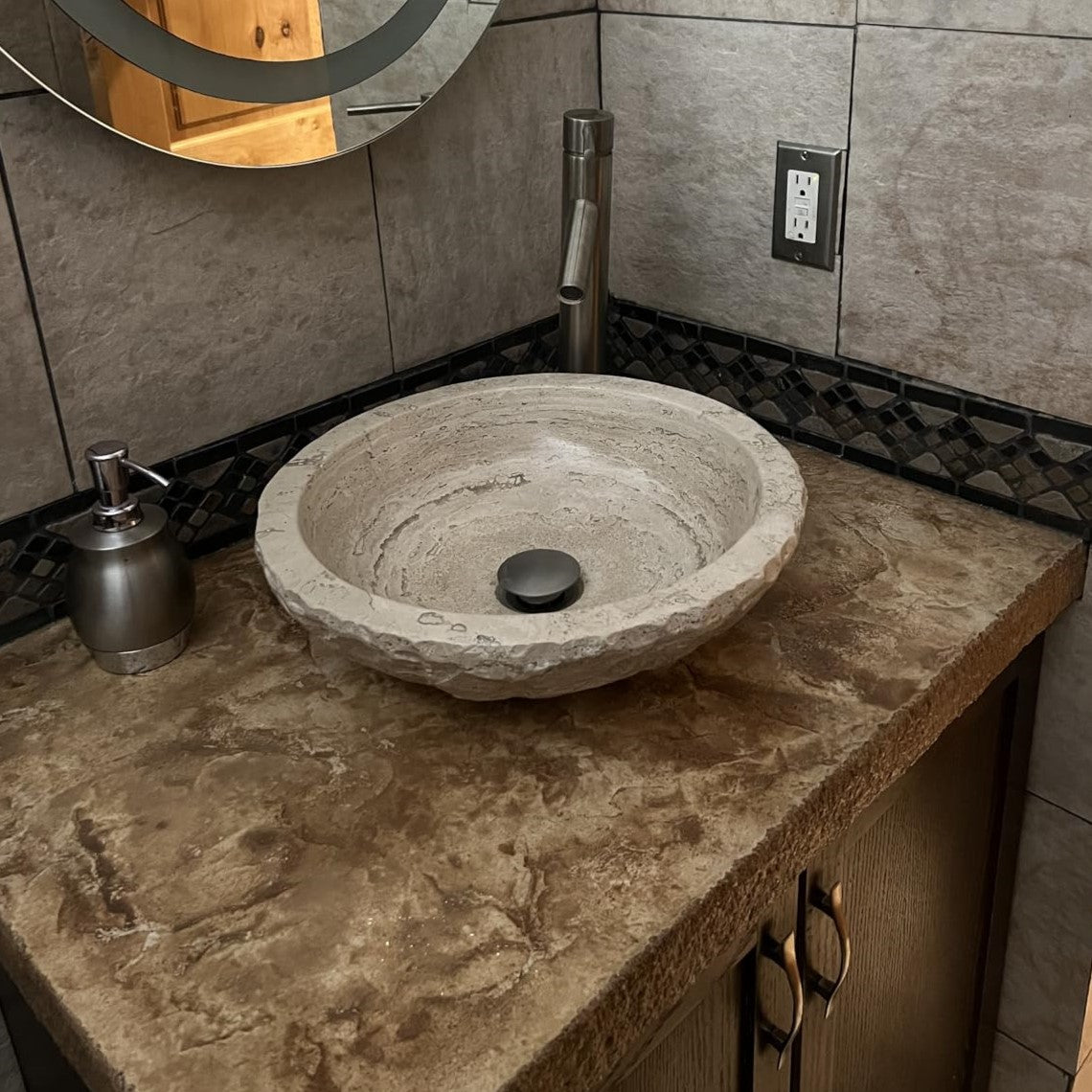 Round beige travertine vessel sink with a hand-carved chiseled exterior, styled on a rustic brown stone countertop with a brushed nickel faucet and matching soap dispenser in a bathroom setting.