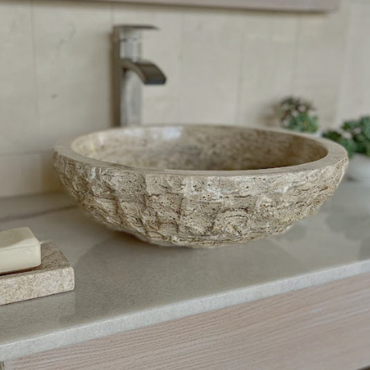 Round beige travertine natural stone vessel sink with a rustic chiseled exterior and smooth interior basin, styled with a polished soap tray on a  vanity countertop.