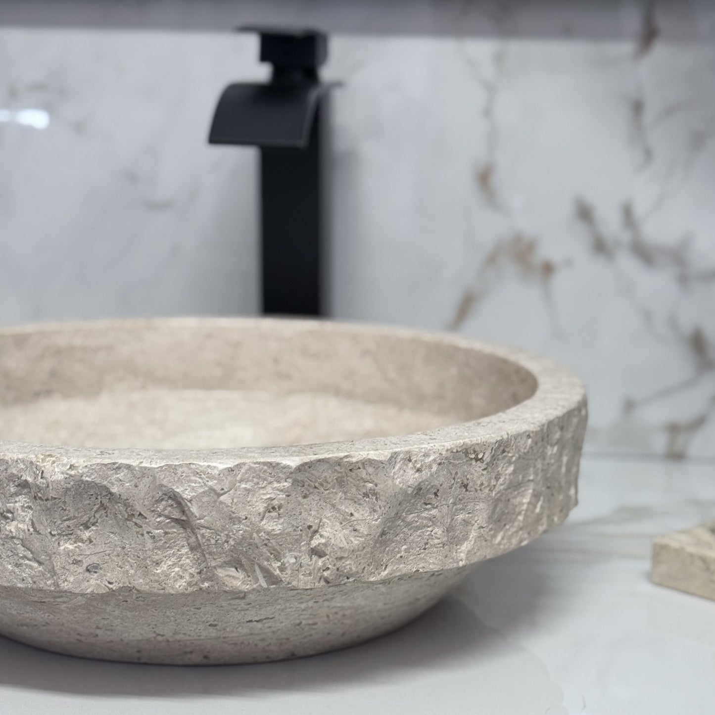 Handmade tan travertine bathroom wash basin featuring a rustic textured chiseled border and a sleek polished lower base, styled with a black faucet backdrop.