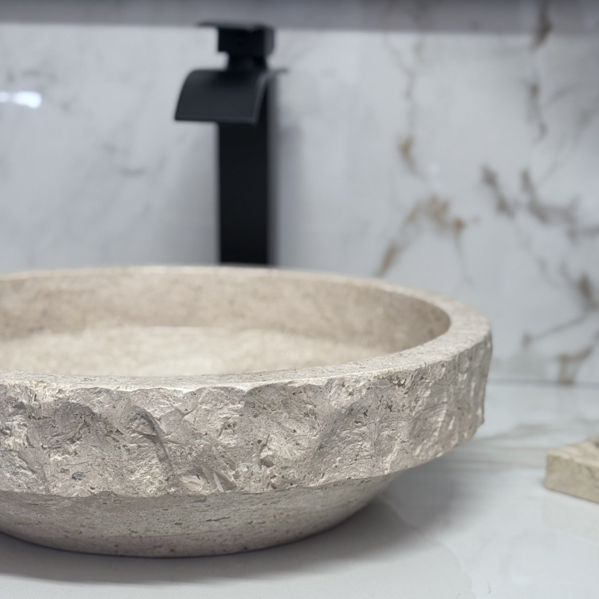 Handmade tan travertine bathroom wash basin featuring a rustic textured chiseled border and a sleek polished lower base, styled with a black faucet backdrop.