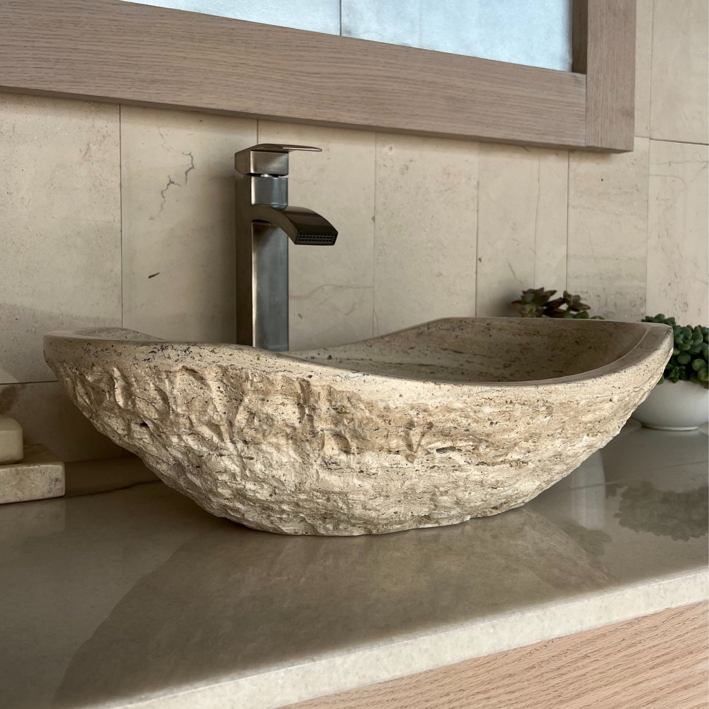 Side-angle profile of a rustic beige travertine bathroom basin, highlighting its heavy chiseled outer surface and curved oval silhouette resting on a polished vanity countertop.