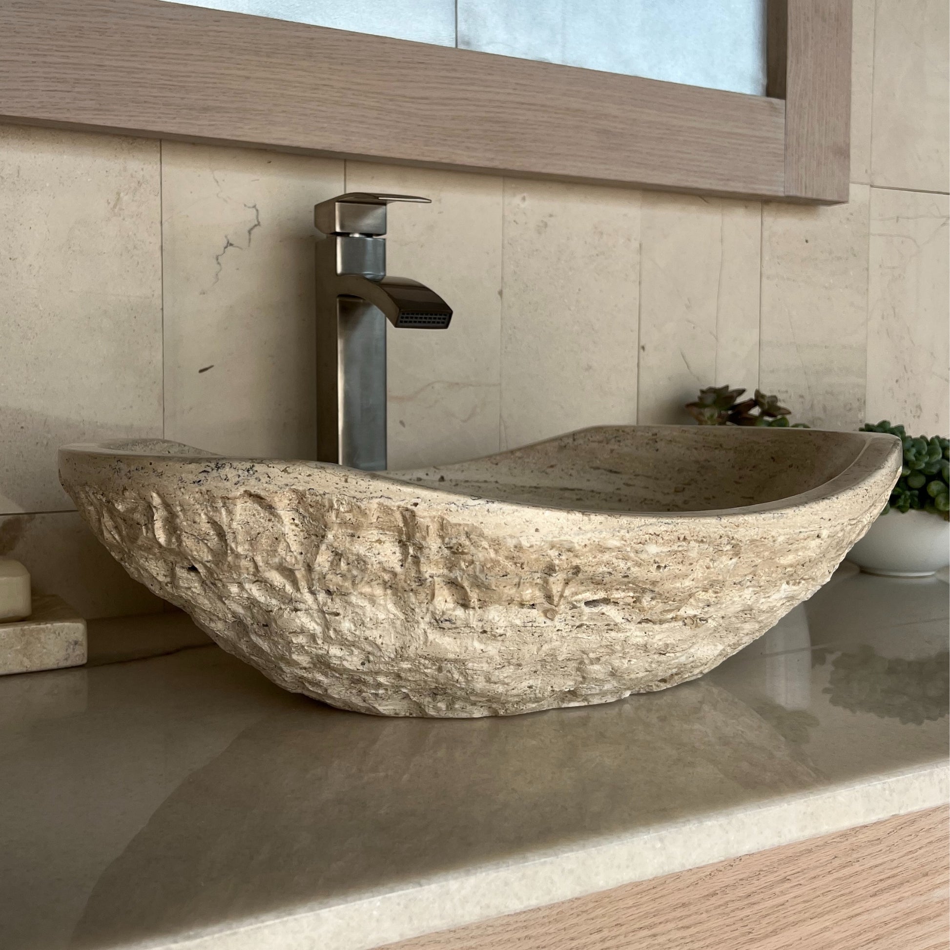 Side-angle profile of a rustic beige travertine bathroom basin, highlighting its heavy chiseled outer surface and curved oval silhouette resting on a polished vanity countertop.