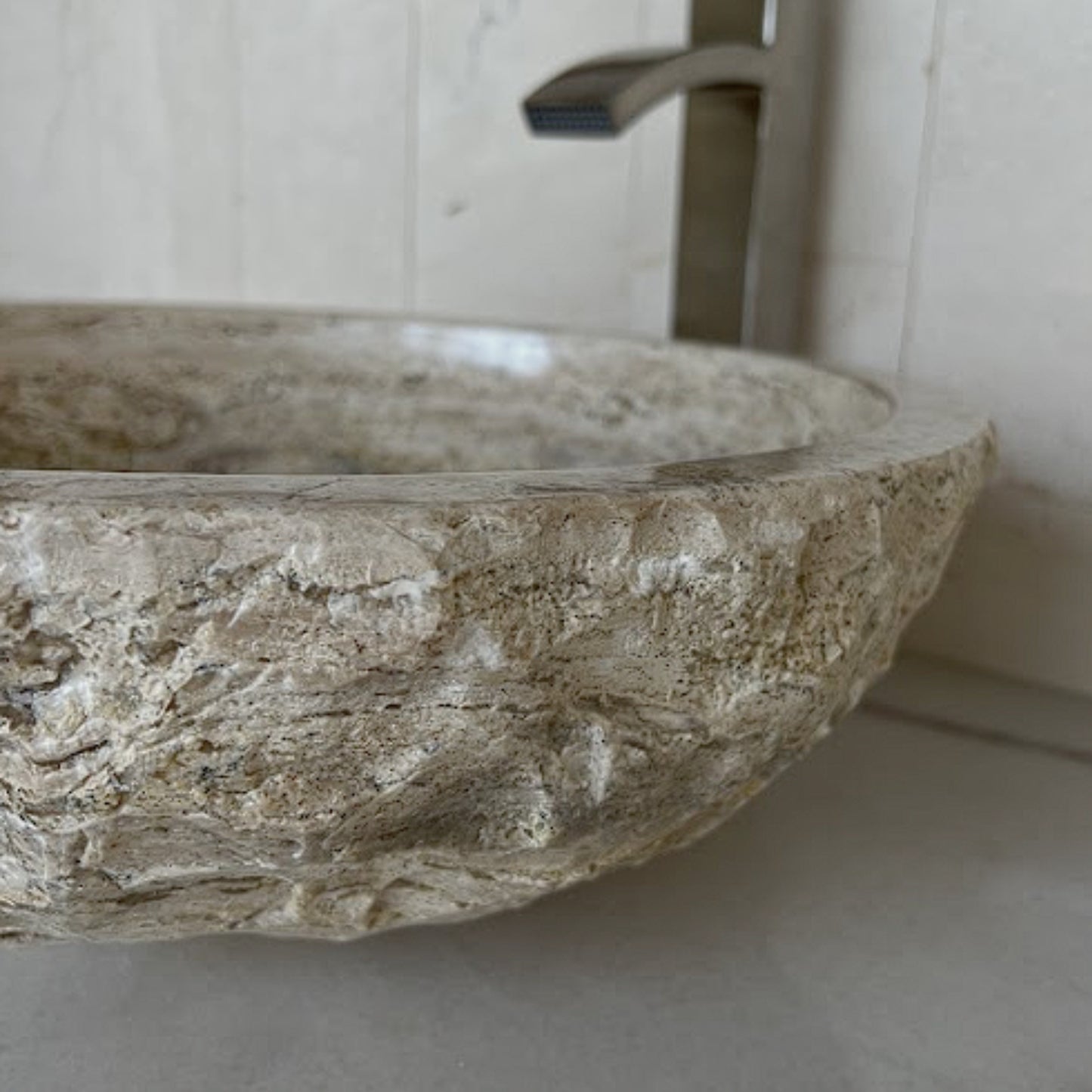 Side view of a round beige travertine vessel sink highlighting the highly textured, hand-carved chiseled exterior and smooth polished inner basin, styled with a modern brushed nickel faucet in the background.