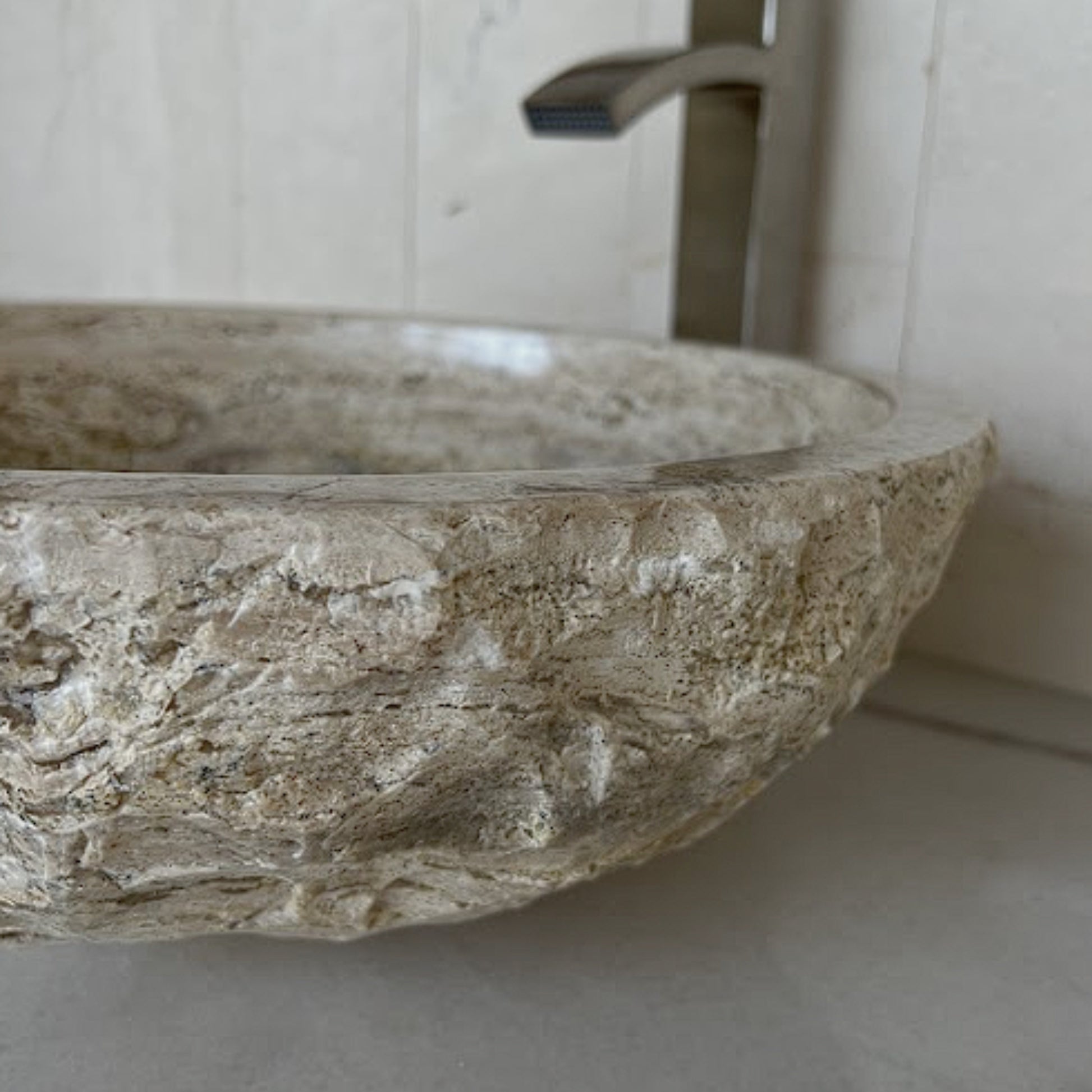 Side view of a round beige travertine vessel sink highlighting the highly textured, hand-carved chiseled exterior and smooth polished inner basin, styled with a modern brushed nickel faucet in the background.