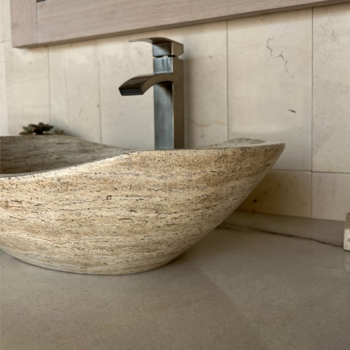 Close-up side view of an oval polished tan travertine vessel sink highlighting the fluid, curved silhouette and smooth natural stone exterior, styled with a modern brushed nickel faucet.