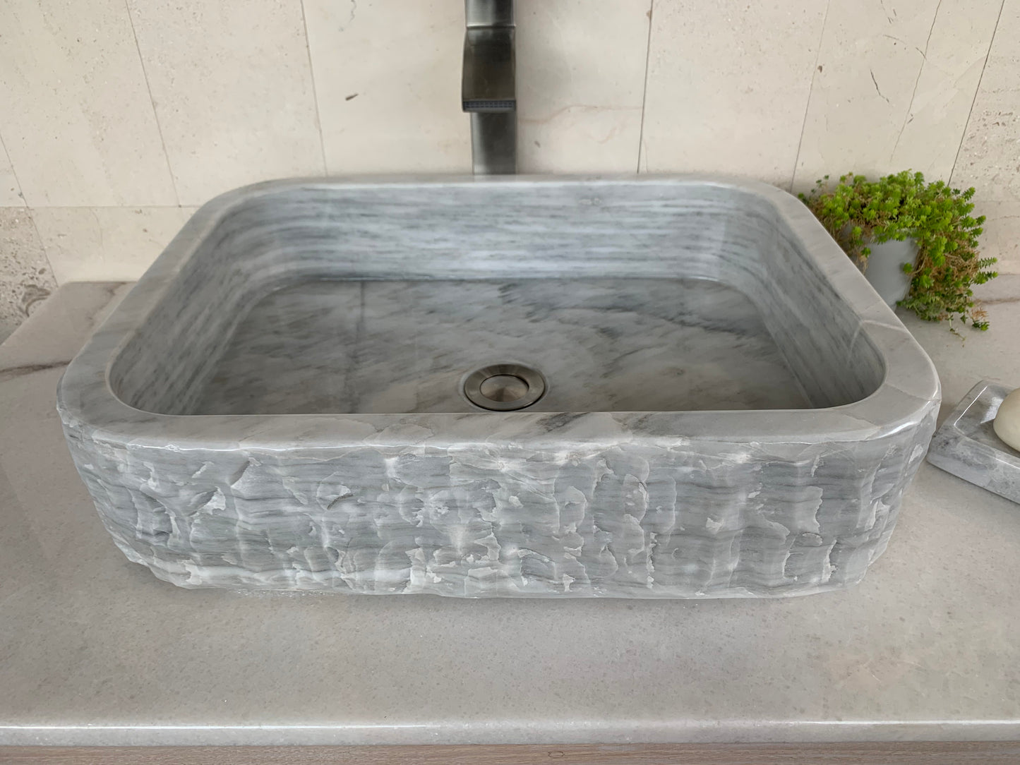 Angled top-down view of a rectangular gray marble bathroom basin, highlighting its polished inner bowl with a central chrome drain fitting, contrasted against a textured chiseled outer surface.