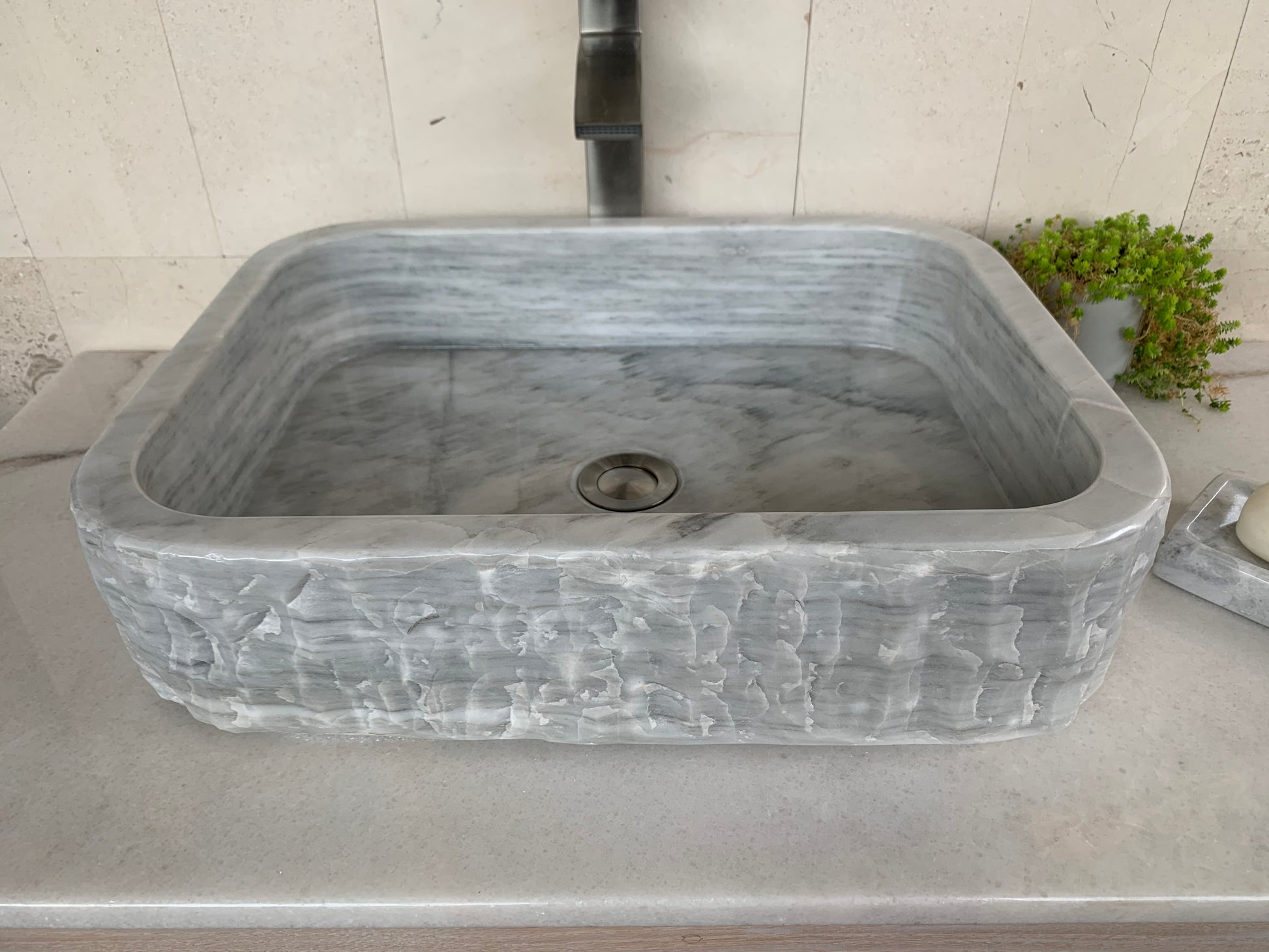Angled top-down view of a rectangular gray marble bathroom basin, highlighting its polished inner bowl with a central chrome drain fitting, contrasted against a textured chiseled outer surface.